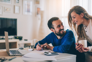 Couple smiling at each other as man sits at table in home working and woman leans over him holding coffee.